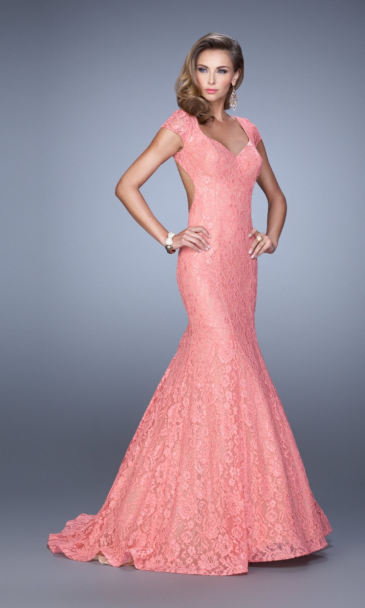 A woman models the coral La Femme 21509, a cap-sleeve long lace prom dress with open back and slight mermaid train, against a plain gray background.